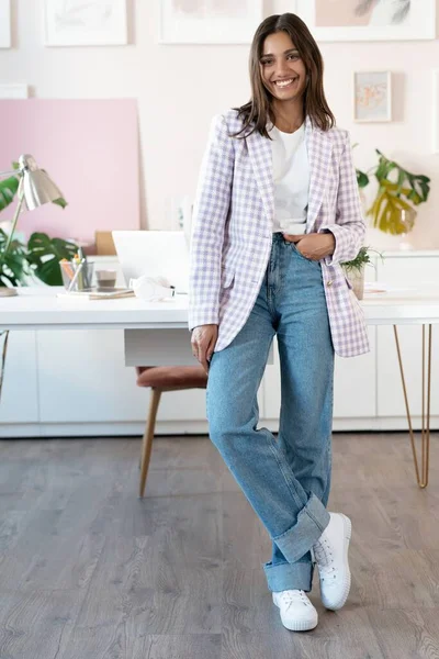 Mujer joven confiada en ropa casual inteligente sonriendo mientras está de pie cerca de su lugar de trabajo — Foto de Stock