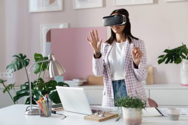 Sanal gerçeklikteki kendine güvenen genç kadın ofisteki çalışma yerinde dururken havaya işaret ediyor.