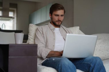 Dizüstü bilgisayar kullanan, evdeki koltukta oturan, sohbet eden ya da internetten alışveriş yapan serbest çalışan bir adam.