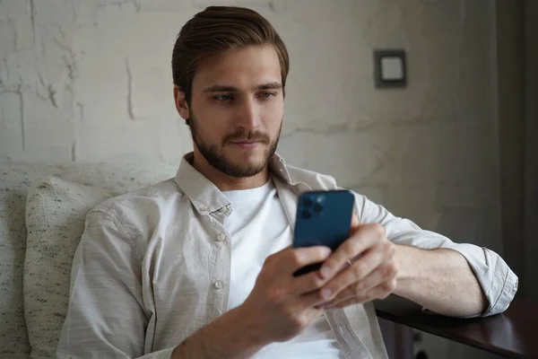 Beau homme en t-shirt basique souriant et tenant le téléphone portable dans les mains tout en étant assis sur le canapé dans le salon. — Photo