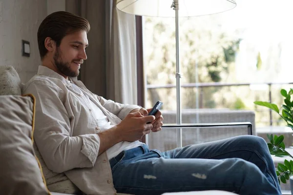 Homem bonito na camiseta básica sorrindo e segurando o telefone celular nas mãos enquanto sentado no sofá na sala de estar. — Fotografia de Stock