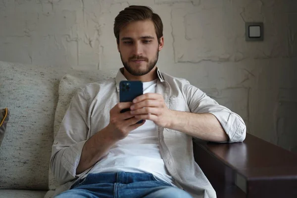 Knappe man in basic t-shirt glimlachend en mobiele telefoon in de hand terwijl hij op de bank zit in de woonkamer. — Stockfoto