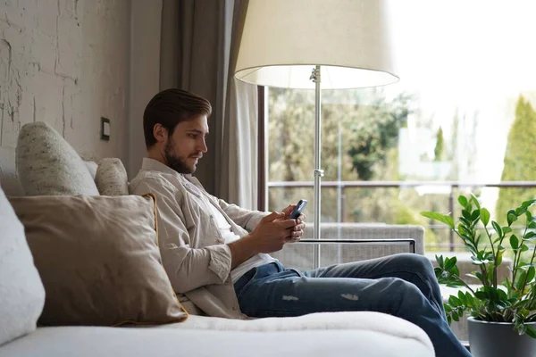 기본 티셔츠를 입고 거실에서 소파에 앉아 미소를 짓고 휴대 전화를 들고 휴대폰을 들고 있는 남자. — 스톡 사진