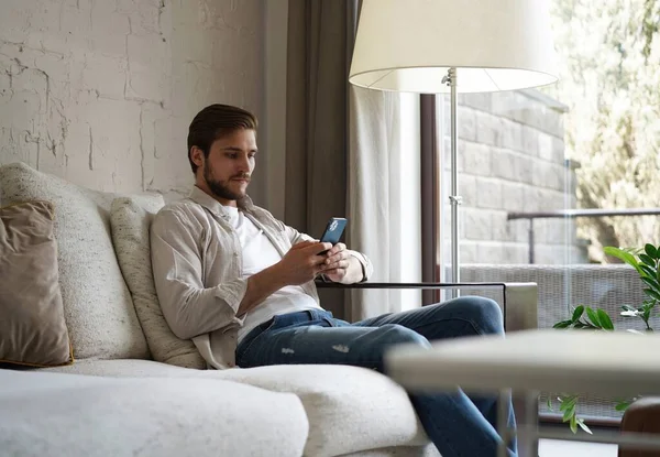Jóképű férfi alapvető pólóban mosolyog és mobiltelefont tart a kezében, miközben a kanapén ül a nappaliban.. — Stock Fotó