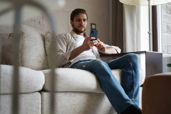 Beau homme en t-shirt basique souriant et tenant le téléphone portable dans les mains tout en étant assis sur le canapé dans le salon. — Photo