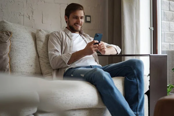 Knappe man in basic t-shirt glimlachend en mobiele telefoon in de hand terwijl hij op de bank zit in de woonkamer. — Stockfoto