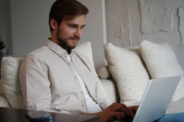 Άντρας που χρησιμοποιεί φορητό υπολογιστή, κάθεται στον καναπέ στο σπίτι, κουβεντιάζοντας ή ψωνίζοντας στο διαδίκτυο, ελεύθερος επαγγελματίας που εργάζονται σε απευθείας σύνδεση — Φωτογραφία Αρχείου