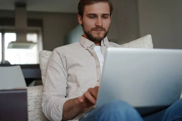 Uomo che utilizza il computer portatile, seduto sul divano a casa, chatta o fa shopping in internet, freelance che lavora online — Foto Stock