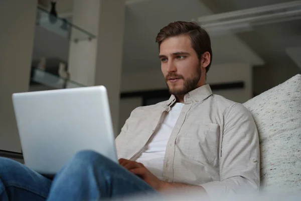 Mann nutzt Laptop, sitzt zu Hause auf Couch, chattet oder kauft im Internet ein, Freiberufler arbeitet online — Stockfoto