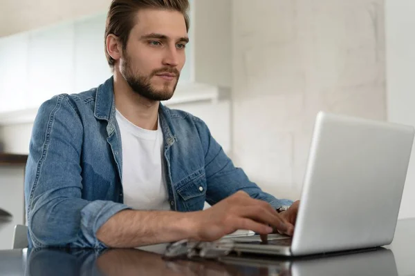 Giovane uomo casuale che lavora sul computer portatile online, seduto a tavola in cucina, guardando lo schermo del computer — Foto Stock