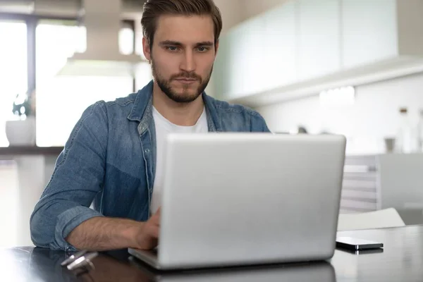 Νέοι casual άνθρωπος που εργάζονται σε φορητό υπολογιστή σε απευθείας σύνδεση, κάθεται στο τραπέζι στην κουζίνα, κοιτάζοντας οθόνη υπολογιστή — Φωτογραφία Αρχείου