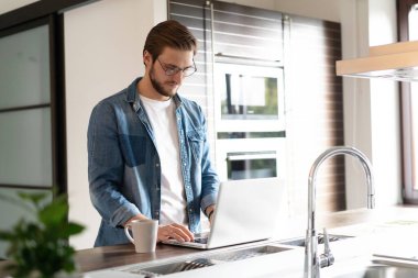 İnternette dizüstü bilgisayarda çalışan genç bir adam mutfaktaki masanın yanında duruyor ve bilgisayar ekranına bakıyor.