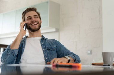 Gülümseyen adam, bir masada oturuyor, dizüstü bilgisayarıyla konuşuyor, önünde bir defter var.