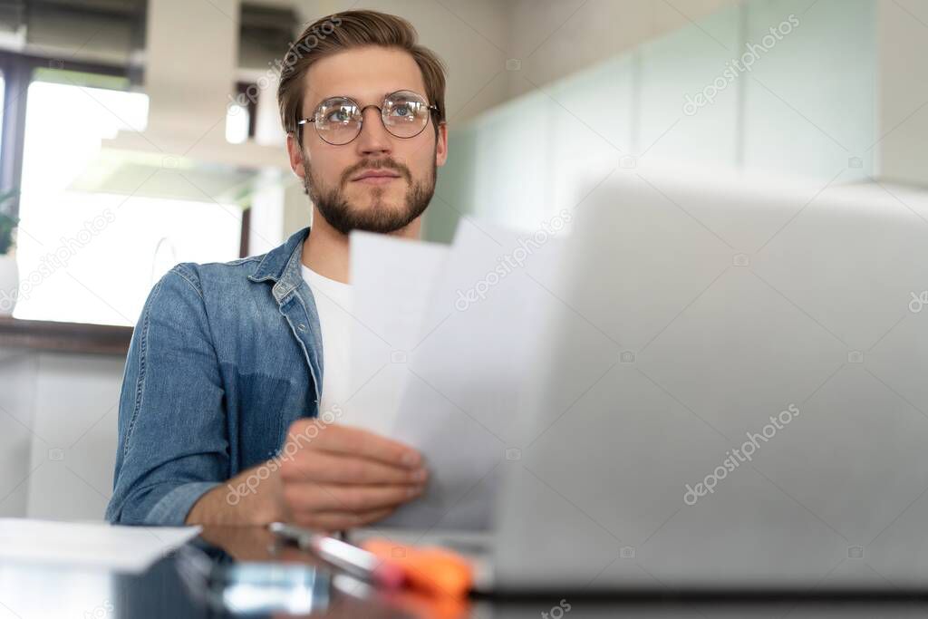 Young man working from home doing paperwork while using laptop.