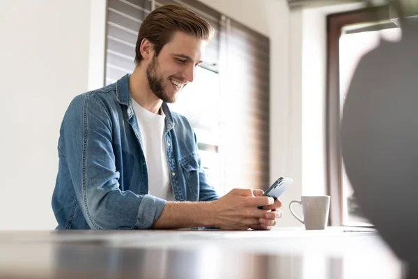 Homme confiant tenant un smartphone, assis dans la cuisine, bavardant en ligne avec des amis ou des collègues, application de navigation — Photo