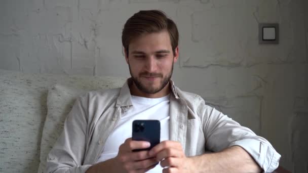 Молодий чоловік використовує смартфон, сидячи на дивані. Чоловік переглядає Інтернет, дивиться відео та використовує соціальні мережі — стокове відео