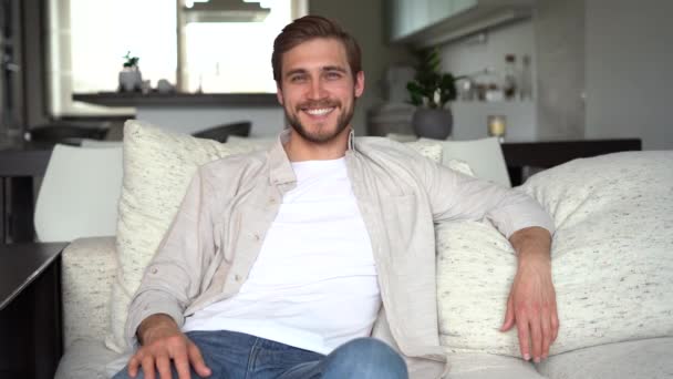 Retrato de un joven feliz sonriendo en casa, relajándose en el sofá. — Vídeos de Stock