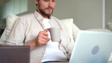 Young adult businessman working in living room with coffee and laptop on sofa. Male freelancer working remotely online