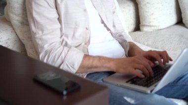 Young man talking on smartphone while sitting on couch with opened laptop, working remotely at home office.