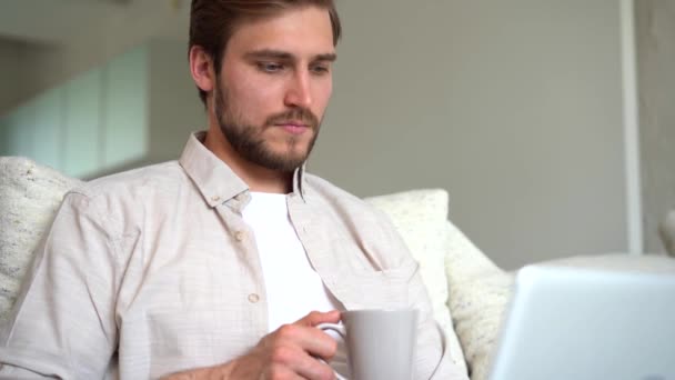 Young adult businessman working in living room with coffee and laptop on sofa. Male freelancer working remotely online — Video Stock