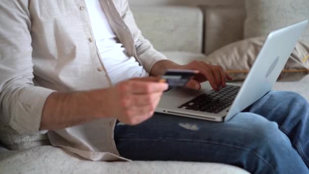 Молодой человек делает онлайн покупки через ноутбук, держит кредитную карту, оплачивает покупки в интернет-магазине — стоковое видео