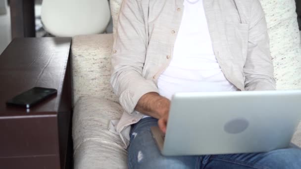 Молодий чоловік розмовляє на смартфоні, сидячи на дивані з відкритим ноутбуком, працює дистанційно в домашньому офісі . — стокове відео