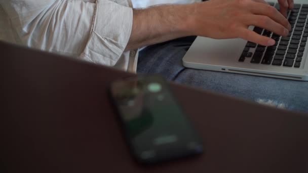 Νεαρός μιλάει στο smartphone ενώ κάθεται στον καναπέ με ανοιχτό φορητό υπολογιστή, που εργάζονται εξ αποστάσεως στο γραφείο στο σπίτι. — Αρχείο Βίντεο