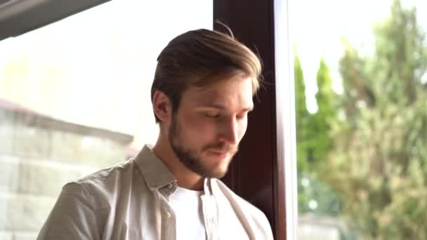 Улыбающийся мужчина в стильной рубашке стоит у окна, используя смартфон — стоковое видео