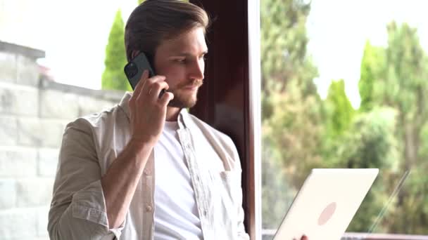 Smiling man in stylish shirt standing near window, using laptop — ストック動画