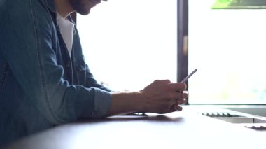 Pencerenin yanında duran gülümseyen adam akıllı telefon kullanıyor. Yakışıklı adam evde modern aletlerle vakit geçiriyor.