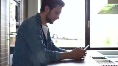 Pencerenin yanında duran gülümseyen adam akıllı telefon kullanıyor. Yakışıklı adam evde modern aletlerle vakit geçiriyor.