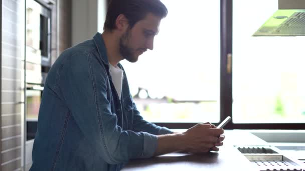 Usmívající se muž stojí u okna a používá smartphone. Hezký člověk tráví volný čas doma s moderní gadget — Stock video