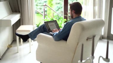 Genç adam koltukta oturur, dizüstü bilgisayar kullanır, çalışır veya uzaktan modern kablosuz teknoloji ve internet bağlantısını kullanarak çalışır.