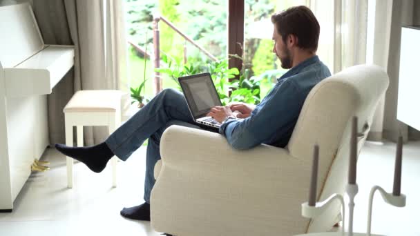 El hombre joven se sienta en el sillón use el ordenador portátil, trabajando o estudiando a distancia usando la tecnología inalámbrica moderna y la conexión a Internet — Vídeo de stock