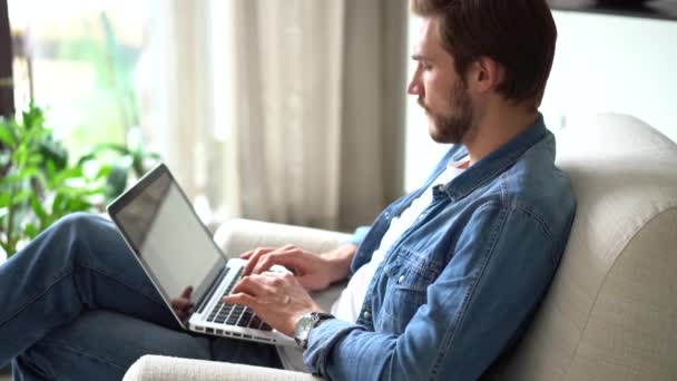 Молодий чоловік сидить у кріслі використовує ноутбук, працює або навчається дистанційно, використовуючи сучасні бездротові технології та підключення до Інтернету — стокове відео