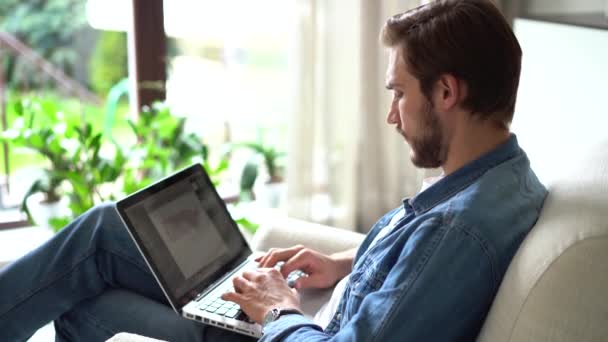 Νεαρός κάθεται σε πολυθρόνα laptop χρήση, εργασίας ή μελέτης χρησιμοποιώντας άμεσα σύγχρονη ασύρματη τεχνολογία και σύνδεση στο internet — Αρχείο Βίντεο