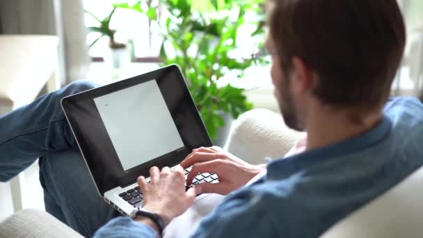 El hombre joven se sienta en el sillón use el ordenador portátil, trabajando o estudiando a distancia usando la tecnología inalámbrica moderna y la conexión a Internet — Vídeos de Stock