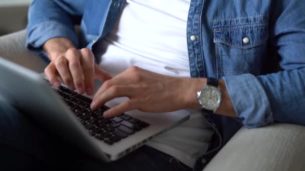 Primer plano de las manos masculinas escribiendo en el portátil. Hombre joven sentado con ordenador portátil en sala de estar, trabajando en interiores — Vídeos de Stock