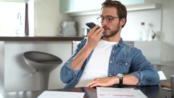 Usmívající se mladý muž nahrává hlasovou zprávu na smartphonu. Fešák pomocí virtuální asistent aplikace sedí doma — Stock video