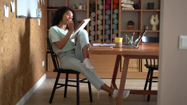 Mladý radostný smíšené-závod žena na volné noze návrhář sedí za stolem, kreslí náčrty v papírové zápisníku — Stock video