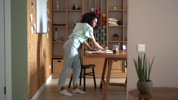 Молодая радостная смешанная раса женщина фрилансер дизайнер, стоящий за столом в офисе на рабочем месте, рисует эскизы в бумажной тетради — стоковое видео