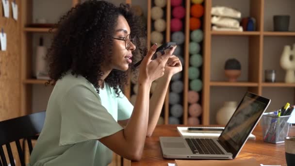 Mujer de raza mixta sentada en el escritorio de la oficina, dictando mensaje de audio en la aplicación móvil, disfrutando de la comunicación a distancia — Vídeo de stock
