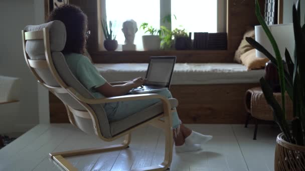 Mujer de raza mixta sentada en un sillón usando una computadora portátil mirando el mensaje de escritura de pantalla, navegando por las redes sociales de Internet — Vídeo de stock