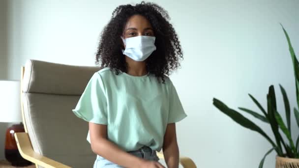 Femme métisse enlevant le masque médical facial protecteur alors qu'elle était assise sur un fauteuil à la maison. — Video