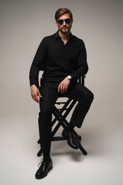 Portrait of a respectable man in a suit and sunglasses sitting on chair on a gray background.