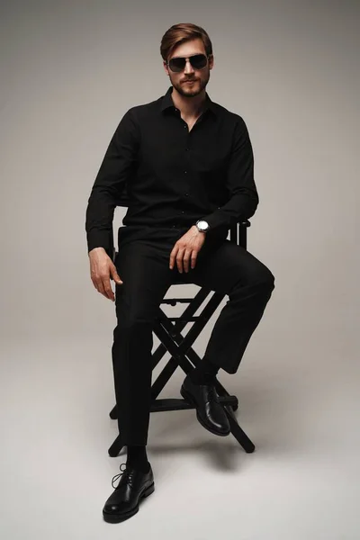 Portrait of a respectable man in a suit and sunglasses sitting on chair on a gray background. — Foto Stock