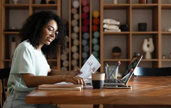 Joven mujer afroamericana hermosa usando su computadora portátil mientras está sentada en la silla en su lugar de trabajo — Foto de Stock