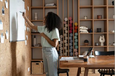 Odaklanmış genç Afro-Amerikan kadın günlük giysiler içinde ofisteki el ilanı çizelgesine yazıyor.