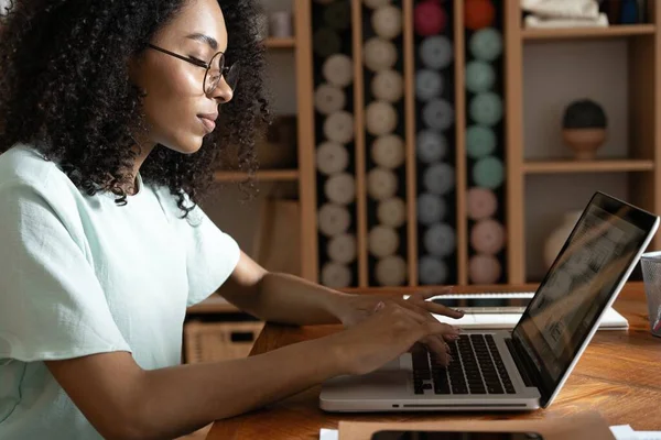 Joven mujer afroamericana hermosa usando su computadora portátil mientras está sentada en la silla en su lugar de trabajo — Foto de Stock