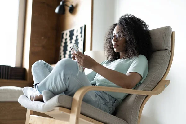 Красива молода змішана раса Жінка використовує смартфон, сидячи на стільці, Серфінг Інтернет, повідомлення в соціальних мережах — стокове фото